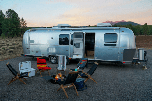 A silver Airstream trailer parked in a scenic area, surrounded by camping chairs and a small fire pit.