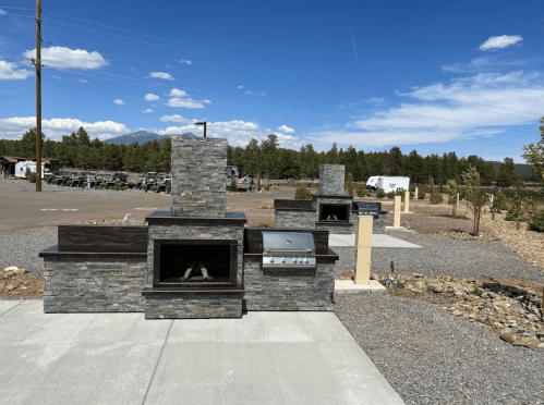 Outdoor kitchen setup featuring a stone grill, fireplace, and countertop, surrounded by gravel and trees.