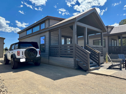A modern cabin with large windows, a porch, and a parked SUV in front, set against a blue sky with clouds.