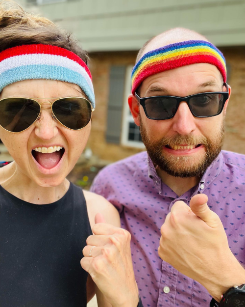 A man and woman wearing colorful headbands and sunglasses, smiling and making excited gestures outdoors.