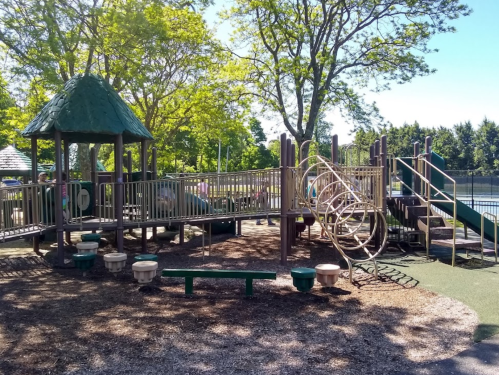 A sunny playground with climbing structures, slides, and benches surrounded by trees.