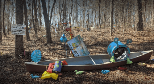 A colorful, whimsical scene in a wooded area featuring a boat surrounded by playful sculptures and signs.