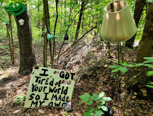 A handmade sign in the woods reads, "I got tired of your world so I made my own," surrounded by trees and a lamp.