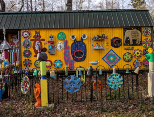 Colorful, whimsical decorations adorn a bright yellow shed, surrounded by a playful fence and vibrant garden art.