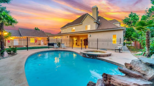 A beautiful backyard with a pool, spa, and a sunset sky behind a two-story house surrounded by palm trees.