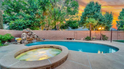 A serene backyard with a pool and hot tub, surrounded by lush greenery and a colorful sunset sky.