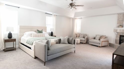 Spacious, modern bedroom with a large bed, cozy seating area, and a fireplace, featuring neutral tones and natural light.