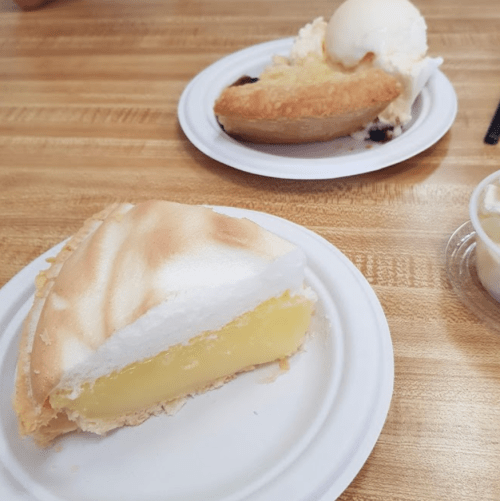 A slice of lemon meringue pie on a white plate, with another dessert and ice cream in the background.