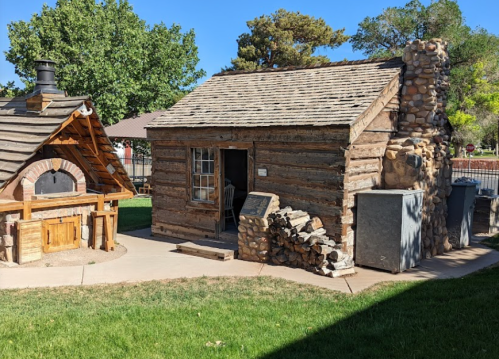 A rustic log cabin with a stone chimney, surrounded by grass and trees, next to an outdoor oven structure.