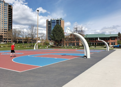 A sunny outdoor basketball court with colorful markings and players shooting hoops in a city park setting.