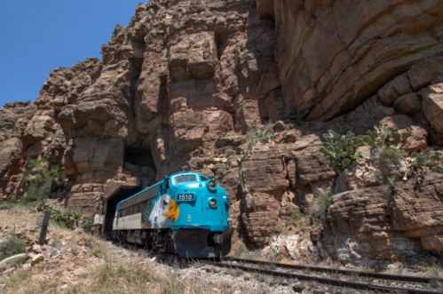 A turquoise train travels along a rocky canyon, surrounded by towering cliffs and desert vegetation under a clear blue sky.