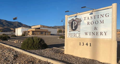 Sign for Pierce Tasting Room & Winery, with a building and mountains in the background under a clear blue sky.