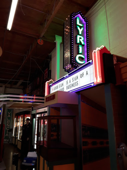 Neon sign for "Lyric" theater with a marquee reading, "A good sign is a sign of a business." Vintage arcade setting.