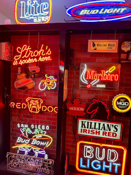 A collection of colorful neon beer and tobacco signs on a brick wall, featuring brands like Bud Light and Marlboro.