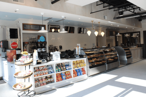 Bright café interior featuring a display case of pastries, snacks, and a coffee station with seating areas.