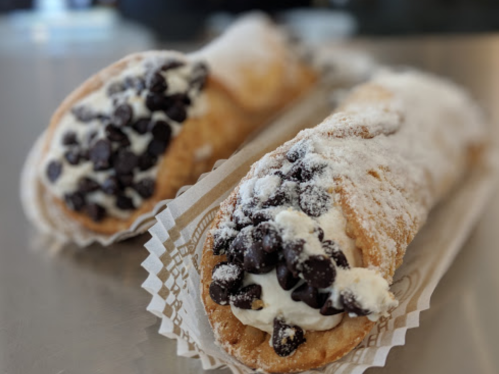 Two cannoli filled with cream and topped with chocolate chips, dusted with powdered sugar on a paper tray.