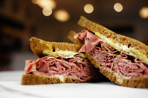 Close-up of a sandwich with layers of sliced meat on dark bread, with mustard and mayonnaise visible.