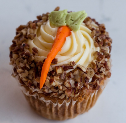 A cupcake topped with cream frosting, a carrot decoration, and chopped pecans around the edges.