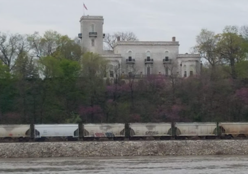 A historic building atop a hill overlooks a river, with freight train cars lined along the water's edge.