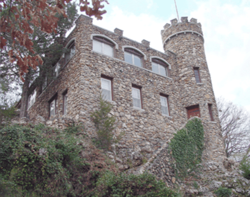 A stone castle with a turret, surrounded by greenery and trees, set against a cloudy sky.