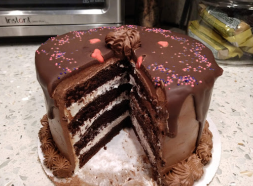 A chocolate layer cake with frosting and sprinkles, partially sliced to reveal its layers.