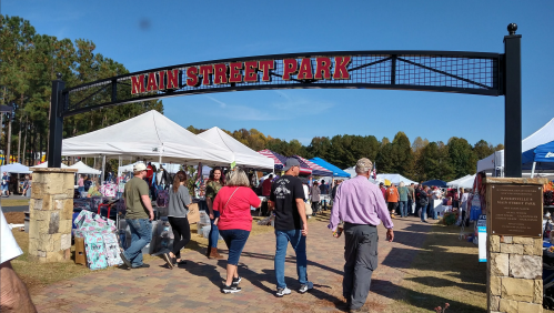 Crowd of people walking through Main Street Park, with tents and stalls set up for a market event on a sunny day.