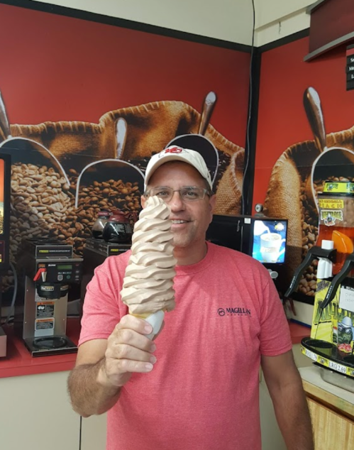 A man in a red shirt holds a large swirl ice cream cone in a shop with coffee-themed decor.