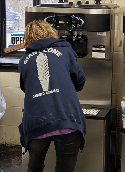 A person in a blue hoodie with "Giant Cone" on the back is using an ice cream machine in a shop.