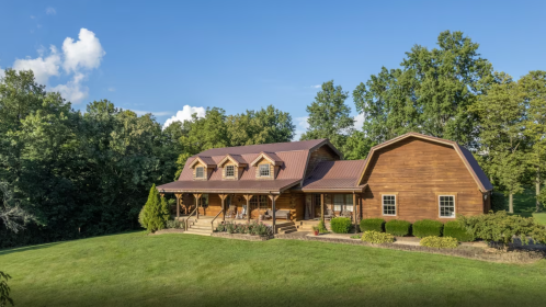 A spacious log cabin surrounded by greenery under a clear blue sky, featuring a large porch and landscaped yard.