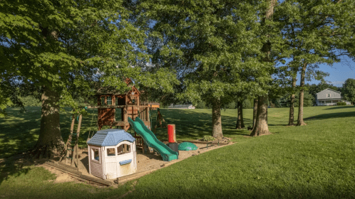 A playground with a wooden playset, slide, and small house, surrounded by trees and a grassy area.
