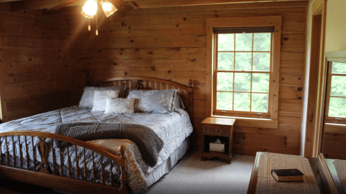 Cozy bedroom with a wooden bed, gray bedding, and a window overlooking greenery. Warm wooden walls and soft lighting.