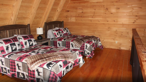 Cozy attic bedroom with two beds, wooden walls, and plaid bedding featuring bears, alongside a nightstand and lamp.