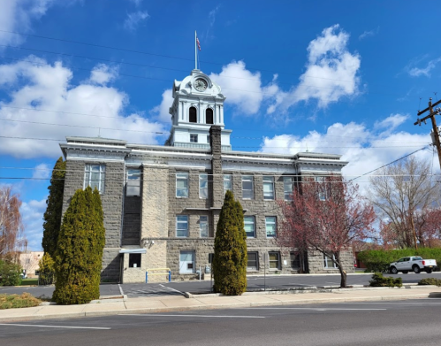 Historic stone building with a clock tower, surrounded by trees and blue sky with clouds.