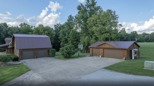 A spacious driveway leads to two wooden garages surrounded by greenery and a basketball hoop in a serene outdoor setting.