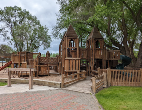 Wooden playground structure with slides and climbing areas, surrounded by trees and grassy areas under a cloudy sky.