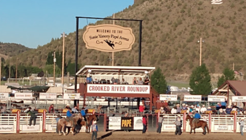 A rodeo arena with spectators, horses, and a sign reading "Welcome to the Susie Yancey Pape Arena" in a scenic setting.