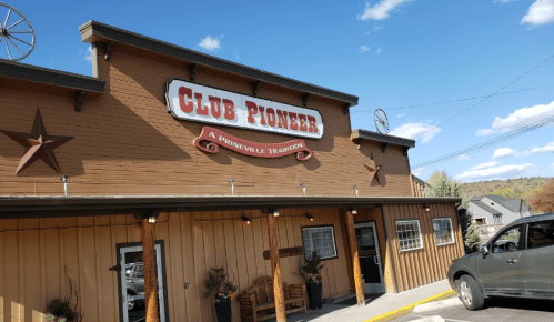 Exterior of Club Pioneer, a rustic building with a sign, surrounded by trees and a clear blue sky.