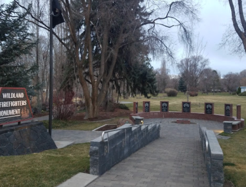 A pathway leads to the Wildland Firefighters Monument, surrounded by trees and memorial plaques in a park setting.