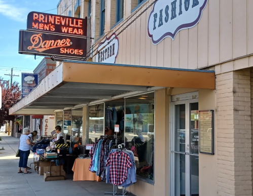 A storefront featuring "Prineville Men's Wear" and "Danner Shoes," with a display of clothing and a customer browsing.