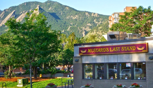 A hot dog stand named "Mustard's Last Stand" with mountains in the background and greenery in the foreground.
