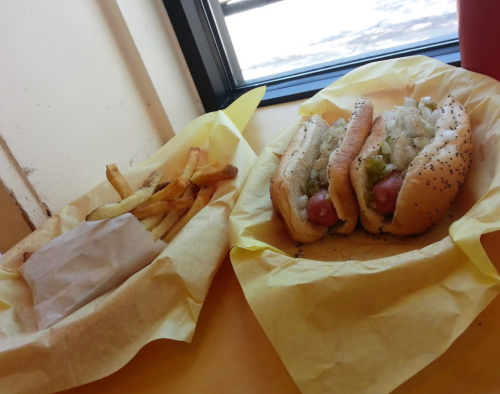 Two hot dogs with toppings in a yellow tray, alongside a serving of fries in a paper wrapper.