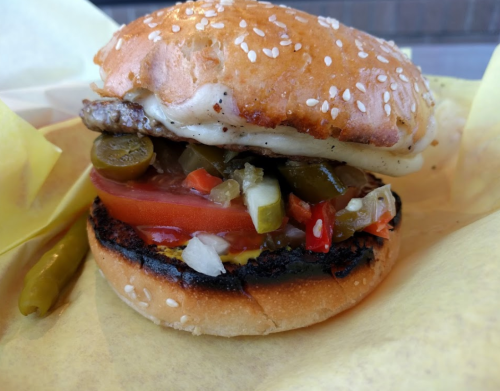 A close-up of a sesame seed burger with a grilled patty, tomatoes, pickles, jalapeños, and mustard on a toasted bun.