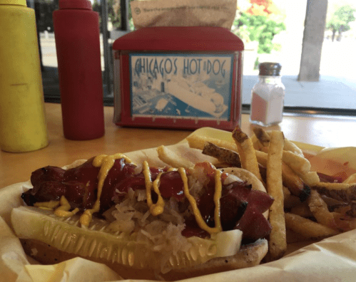 A Chicago-style hot dog with mustard, ketchup, and sauerkraut, served with fries and condiments in the background.