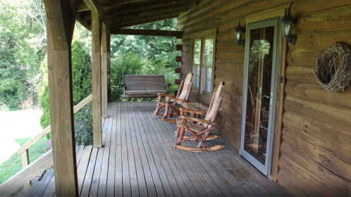 A rustic wooden porch with rocking chairs and a swing, surrounded by greenery and trees.