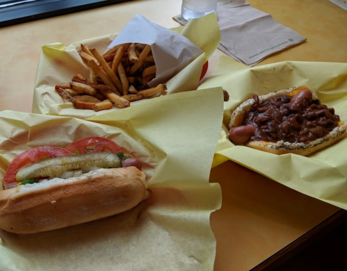 A table with two hot dogs topped with condiments and a side of crispy French fries in yellow paper wrappers.