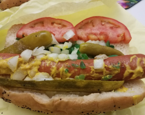 A hot dog in a bun topped with mustard, diced onions, sliced tomatoes, and pickles on a yellow wrapper.