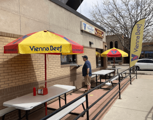 A man stands at a window of a Vienna Beef stand with yellow umbrellas and tables outside, on a sunny day.