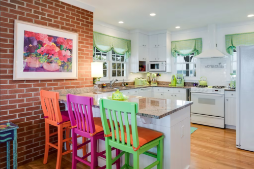Bright kitchen with a brick wall, colorful bar stools, and modern appliances, featuring floral decor and green accents.