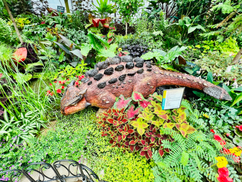 A colorful garden featuring a large, decorative dinosaur sculpture surrounded by vibrant plants and flowers.
