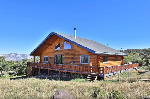 A spacious log cabin with a large deck, surrounded by greenery and mountains under a clear blue sky.